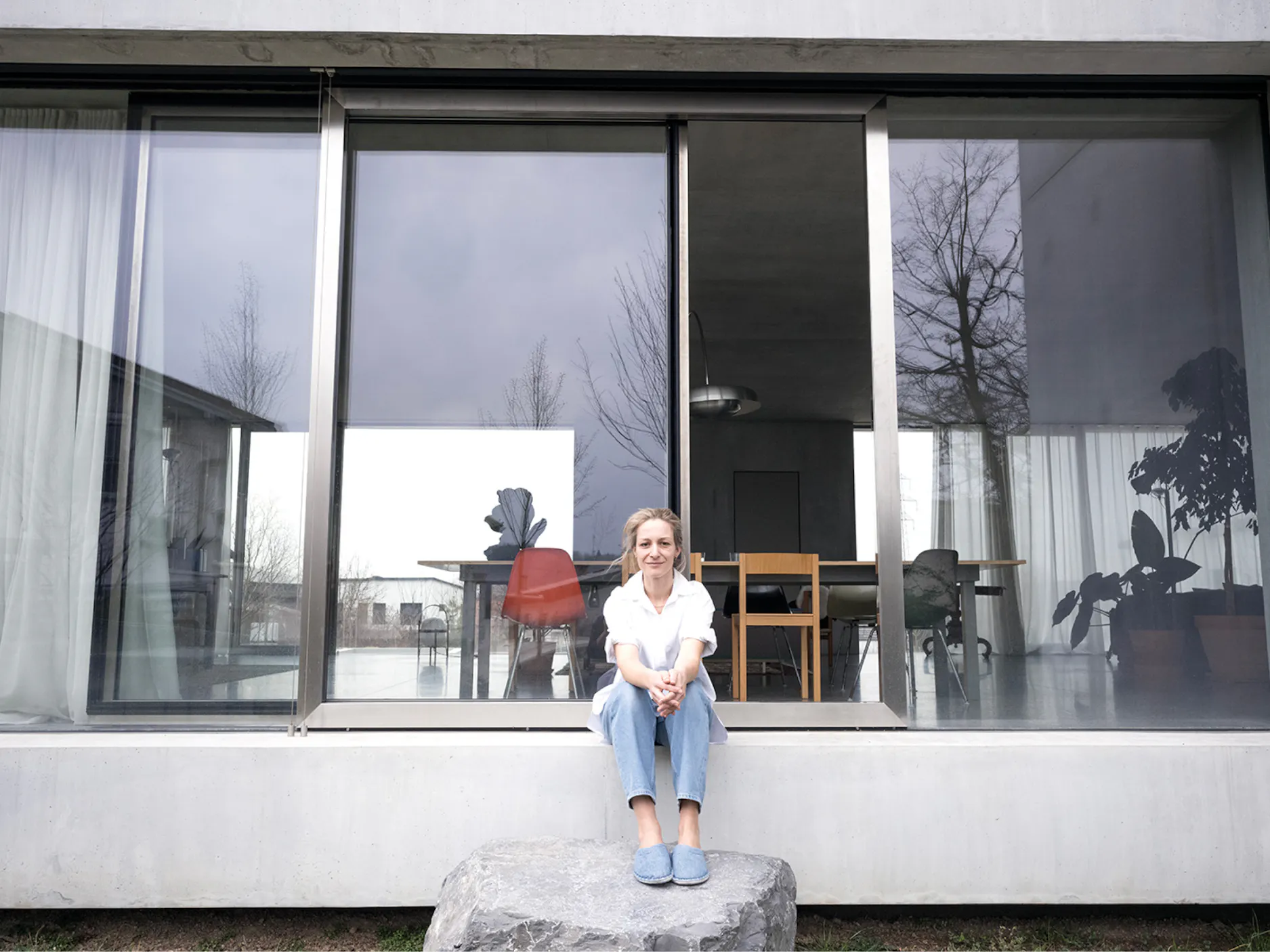 Ženska sedi na vhodu svojega doma. V ozadju so steklena panoramska stekla. Skozi stekla se vidi notranji dnevni prostor z mizo in stoli.